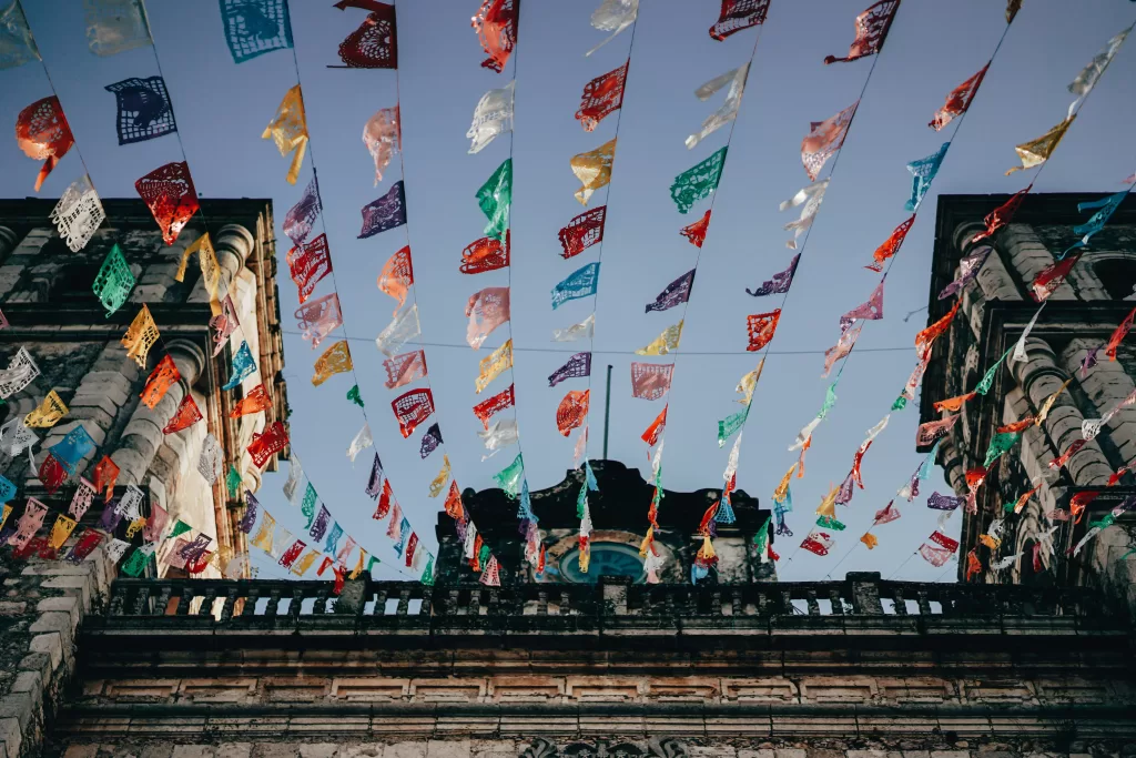 Des guirlandes de papel picado dansent au-dessus d’une architecture ancienne. Une scène festive et haute en couleur.