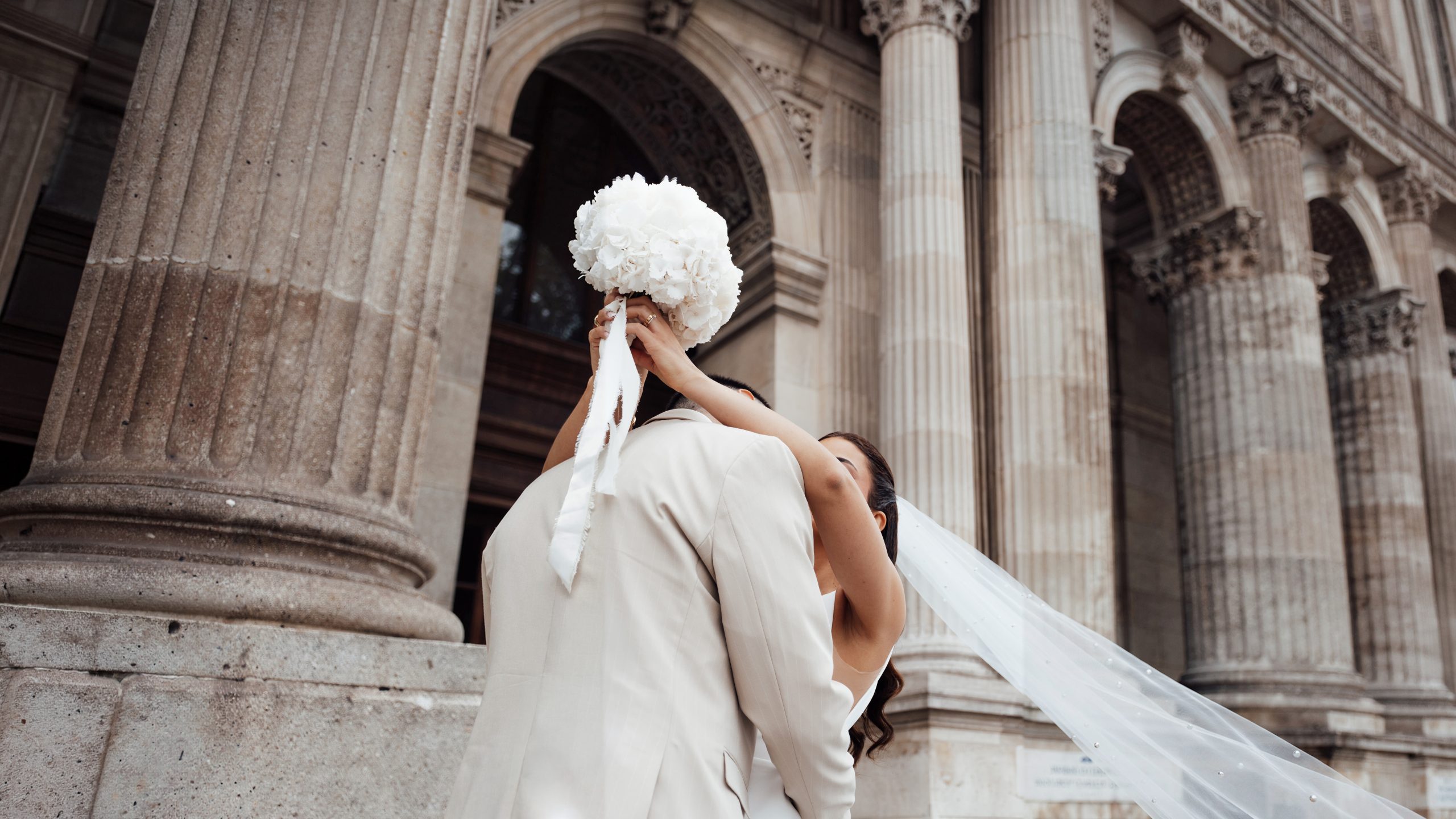 Un couple de jeunes mariés pose devant la façade monumentale d’un édifice en pierre, décorée de grandes colonnes cannelées, d’arches et de sculptures. La mariée tient un imposant bouquet de fleurs blanches devant son visage, tandis que son long voile blanc flotte sur la droite de l’image.