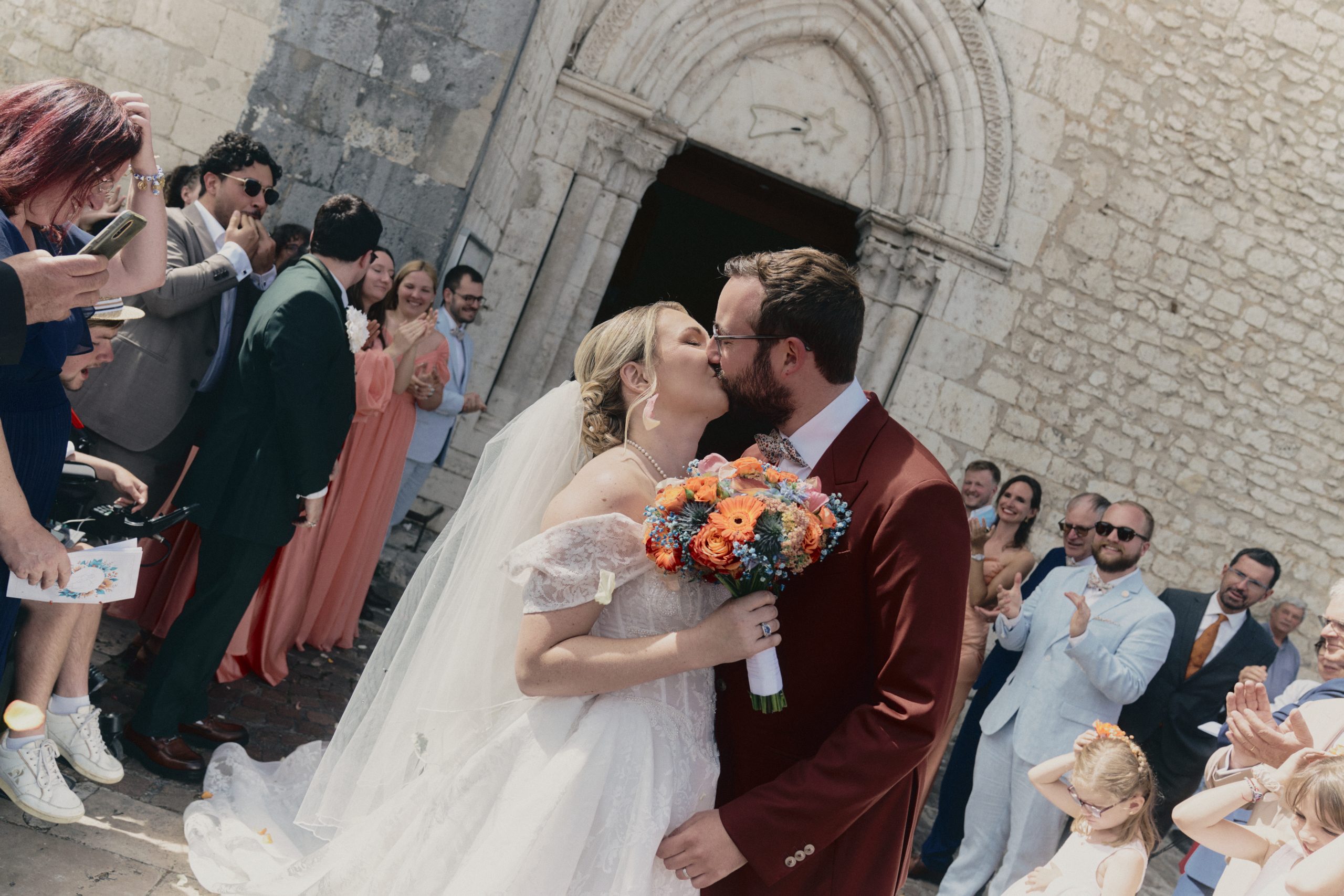 Au premier plan, une mariée en robe blanche et voile embrasse son mari, vêtu d’un costume bordeaux, tout en tenant un bouquet coloré. La scène se déroule devant une ancienne façade en pierre, tandis que leurs invités applaudissent et photographient les jeunes mariés.