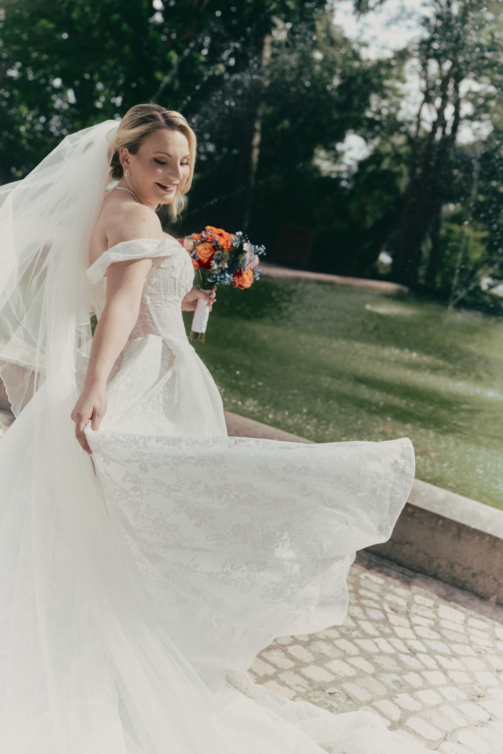 Une mariée blonde sourit en se retournant, vêtue d’une robe blanche en dentelle et d’un long voile. Elle tient un bouquet de fleurs orange, bleues et blanches, tandis qu’elle avance dans un jardin arboré baigné de lumière.