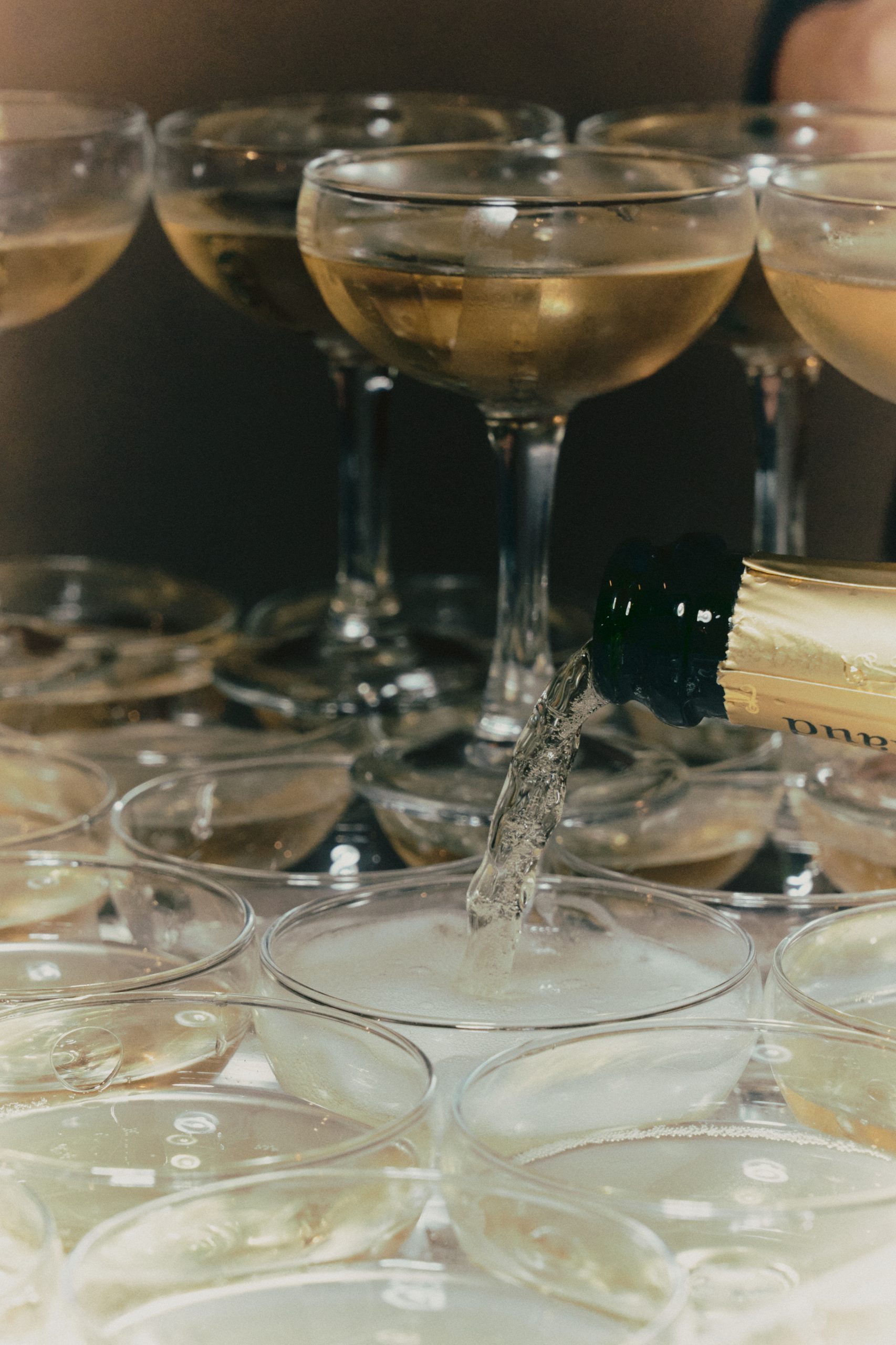 Une bouteille sombre au design travaillé verse un liquide transparent dans un verre placé au premier plan. De nombreux verres se chevauchent autour, créant une composition brillante et chaleureuse dans une ambiance tamisée.