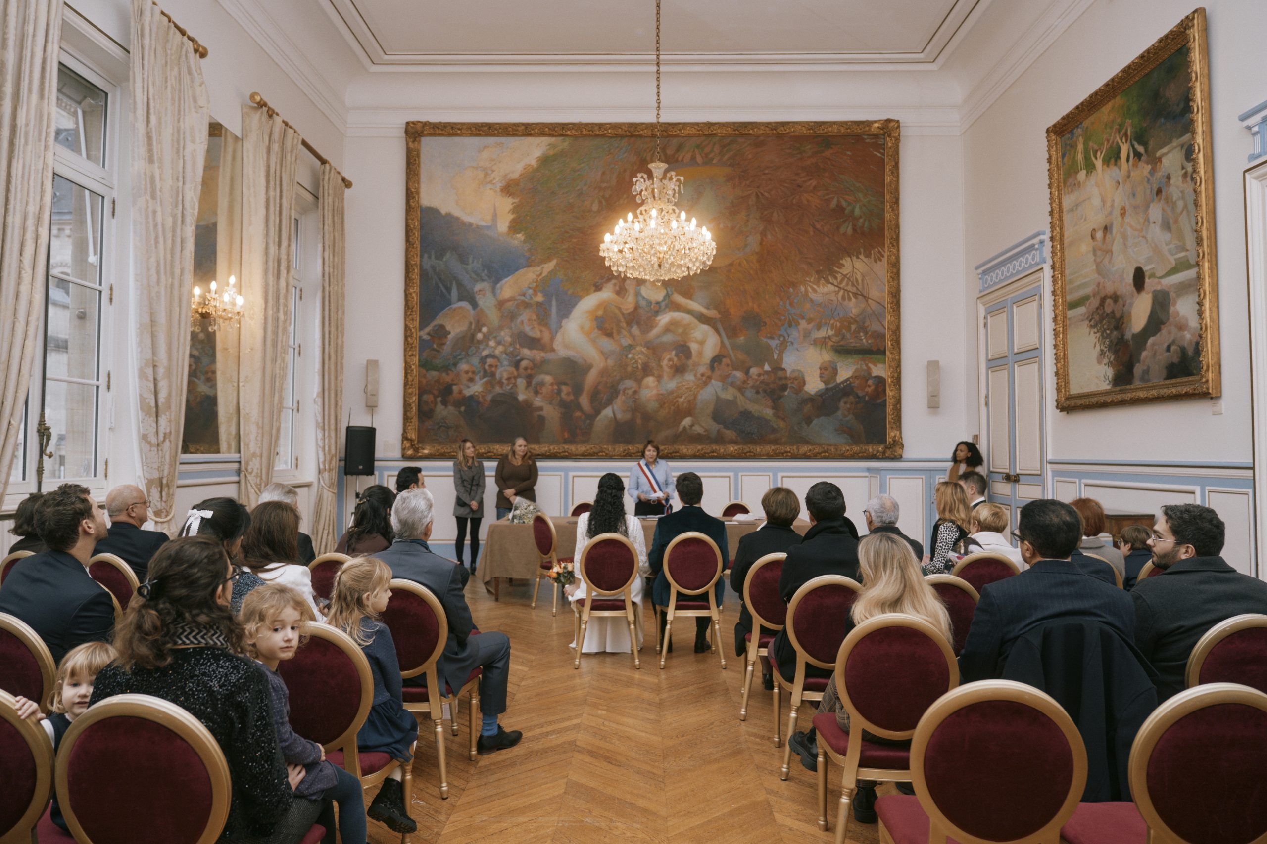 Une assemblée est réunie dans une vaste salle richement décorée, avec parquet en bois, lustre en cristal, moulures et grands tableaux encadrés. Au centre, une femme portant une écharpe tricolore s’adresse aux invités assis, tandis que plusieurs personnes se tiennent derrière elle.