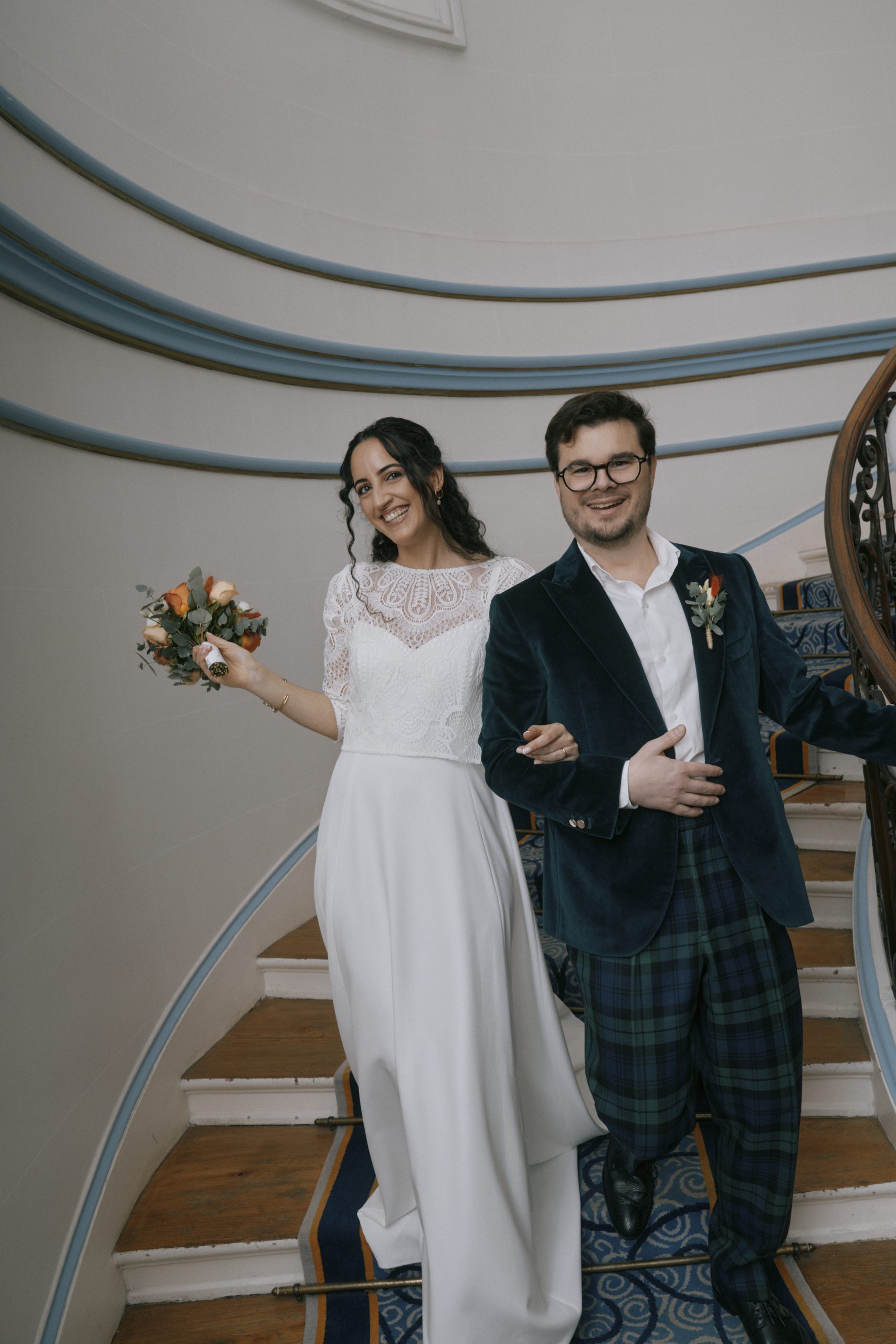 Une mariée vêtue d’une robe blanche en dentelle tient un bouquet de fleurs aux tons pêche et orangés, aux côtés de son mari souriant. Il porte un costume sombre à motif tartan avec une boutonnière fleurie. Le couple pose dans un escalier intérieur courbé, orné de rampes bleu pâle, d’une moquette à motifs et de détails en bois.