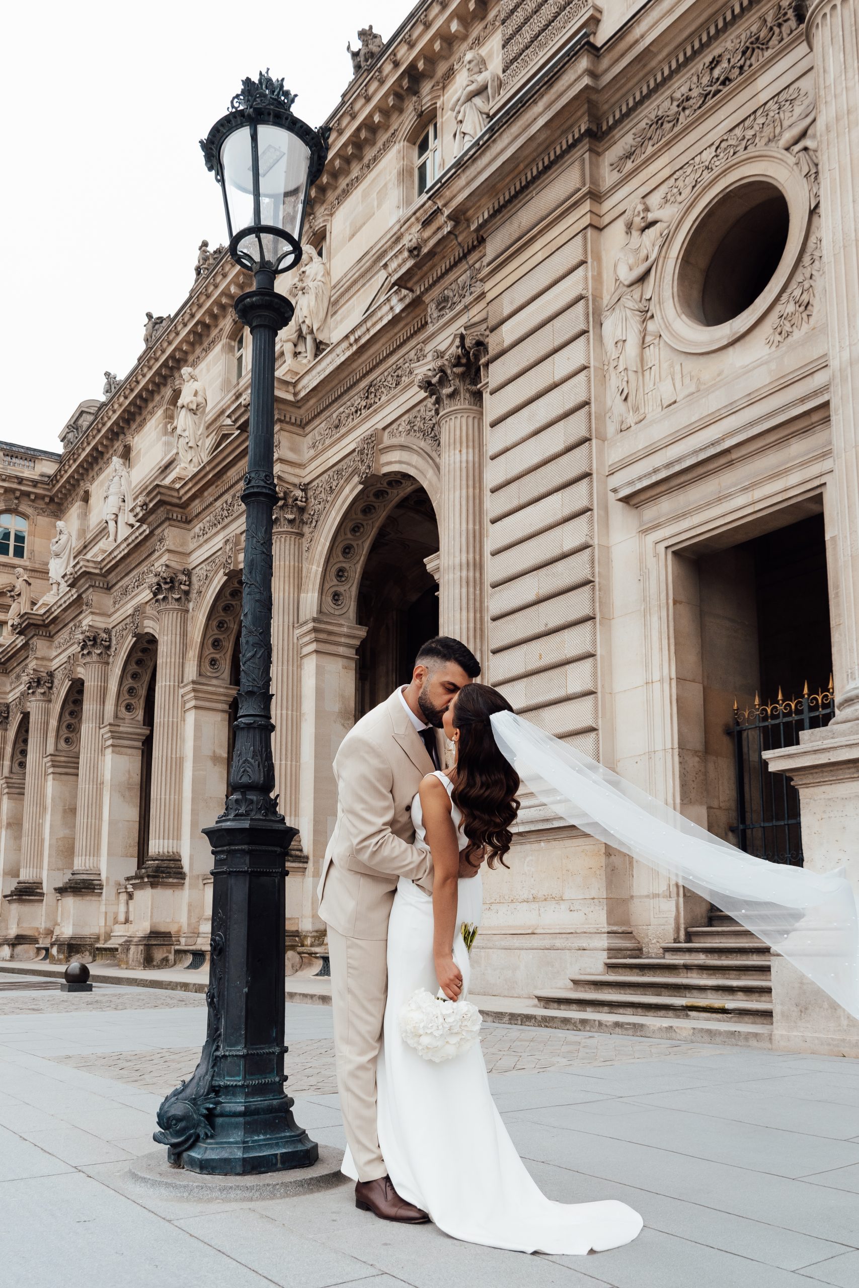 Un couple de jeunes mariés s’embrasse devant un imposant édifice en pierre richement décoré de sculptures, de colonnes et de reliefs architecturaux. Le marié porte un costume clair, tandis que la mariée tient un bouquet blanc et laisse flotter son long voile sur les marches. Un lampadaire noir ouvragé occupe le premier plan.