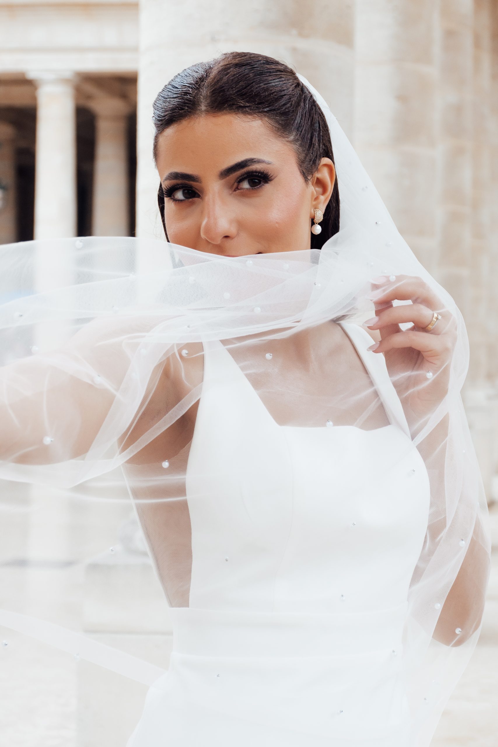 L’image montre en gros plan une mariée au maquillage soigné, aux cheveux foncés attachés et ornés d’un voile blanc. Elle porte des boucles d’oreilles pendantes et une robe blanche en tulle décorée de petites perles, tandis que sa main retient délicatement le voile. L’arrière-plan intérieur, volontairement flou, accentue l’atmosphère intime et lumineuse du portrait.