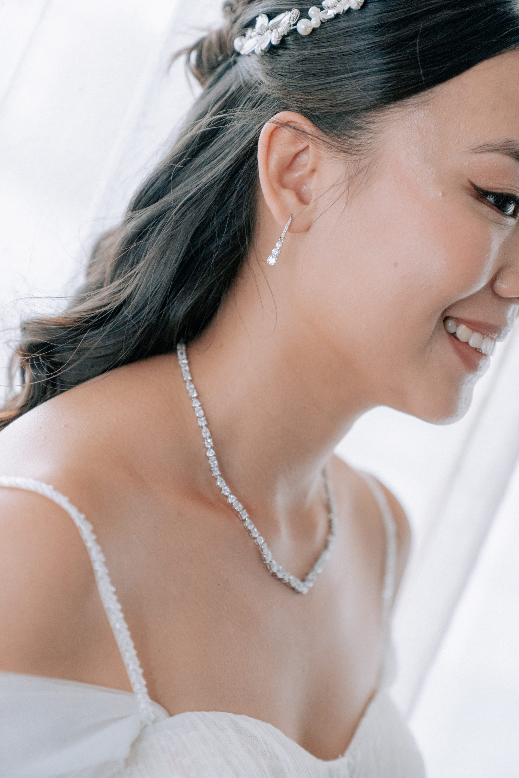 Gros plan du visage souriant d’une mariée aux cheveux foncés, coiffés avec un accessoire orné de perles et de cristaux. Elle porte des boucles d’oreilles pendantes et un collier scintillant, visibles au-dessus de sa robe blanche, dans une lumière douce et lumineuse.