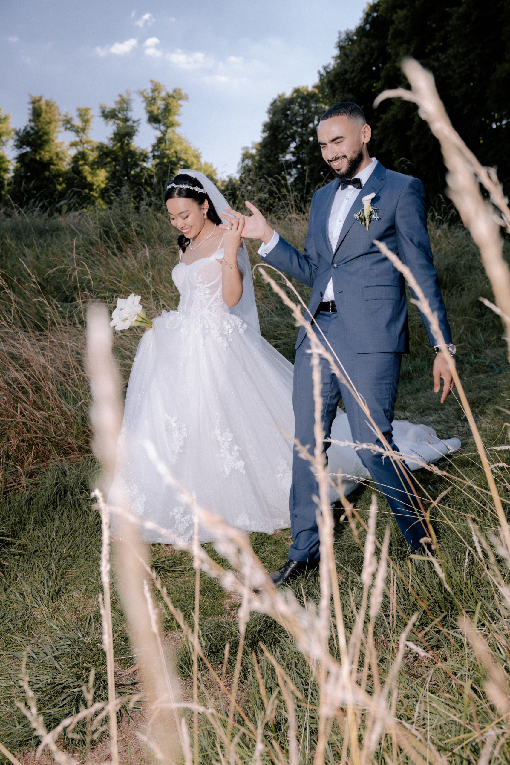 Une mariée en robe blanche et voile marche aux côtés de son époux, vêtu d’un costume bleu avec nœud papillon et boutonnière. Ils avancent dans un champ bordé d’arbres, avec un bouquet blanc à la main et de hautes herbes floues au premier plan.