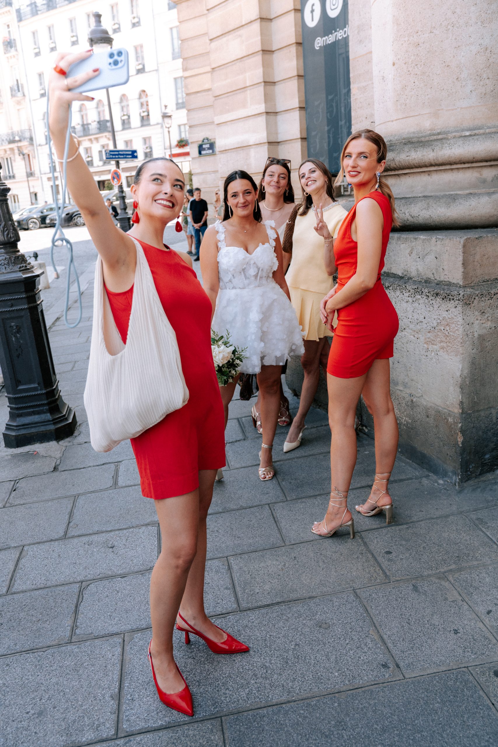 Une femme en robe rouge prend un selfie avec trois amies devant un bâtiment municipal parisien. Au centre, une mariée en robe blanche tient un bouquet, entourée de femmes élégamment vêtues, tandis que l’enseigne de la mairie et les façades urbaines apparaissent en arrière-plan.