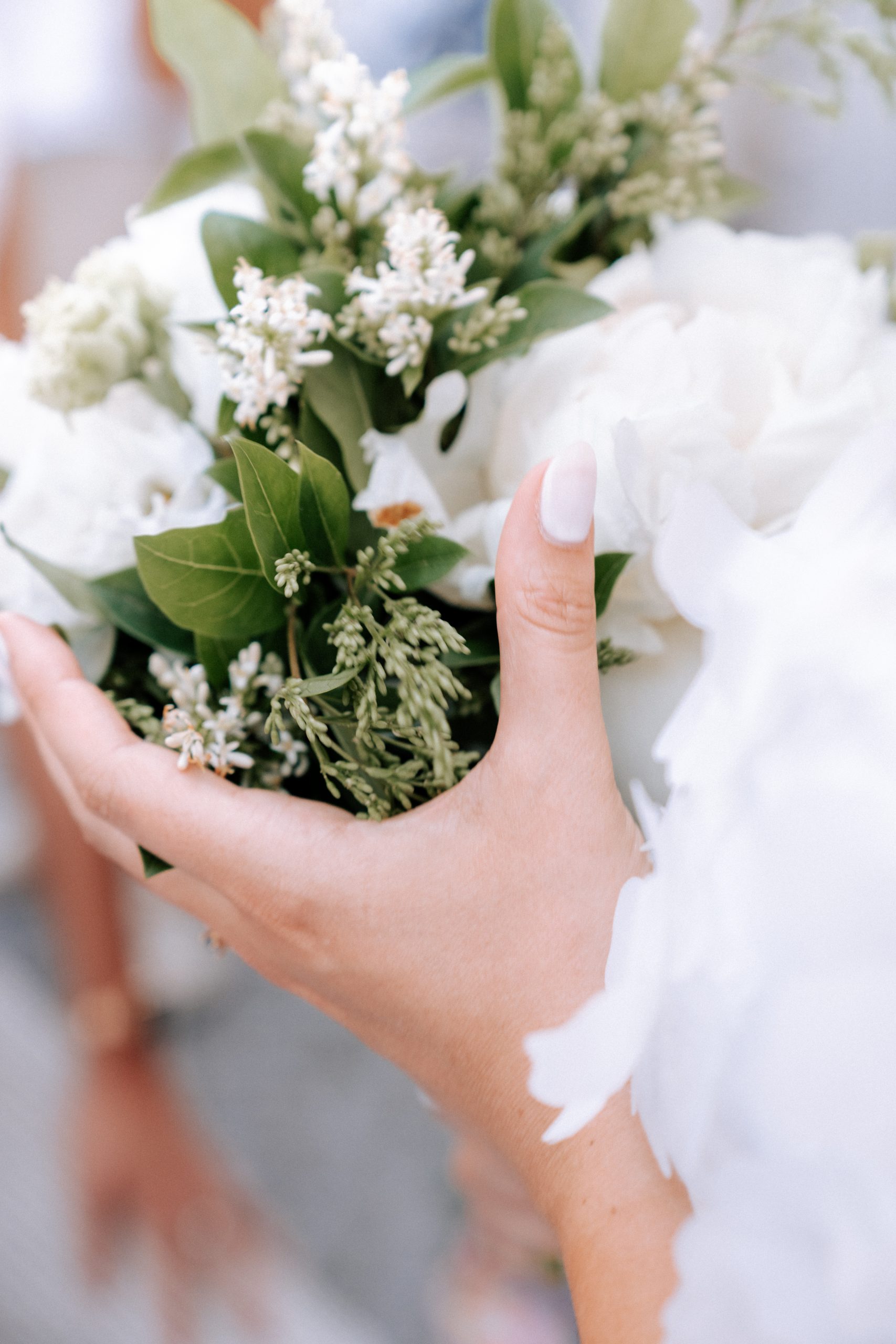 Une personne vêtue de blanc tient entre ses mains un bouquet composé de petites fleurs blanches, de grandes fleurs blanches et de feuillage vert. L’image, photographiée en gros plan avec une faible profondeur de champ, présente une lumière douce et une ambiance romantique.