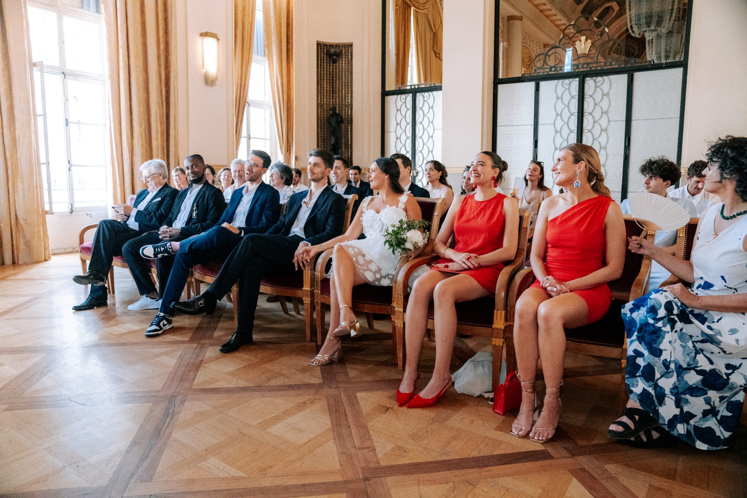 Dans une salle lumineuse aux hauts murs, aux rideaux dorés et au parquet ornementé, plusieurs invités sont assis sur des fauteuils élégants. Au premier plan, une mariée en robe blanche tient un bouquet, entourée de proches vêtus notamment de robes rouges et de costumes sombres.