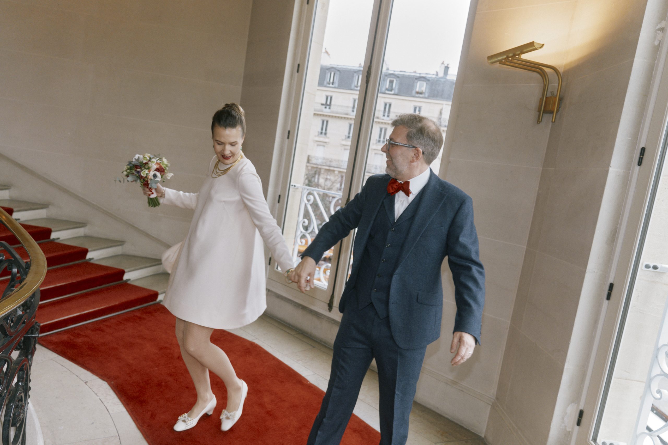 Une mariée en tenue blanche tient un bouquet coloré tandis qu’elle descend un escalier recouvert d’un tapis rouge. À ses côtés, le marié porte un costume bleu avec un nœud papillon rouge et lui tient la main. De grandes fenêtres laissent apparaître les façades élégantes d’un immeuble parisien.