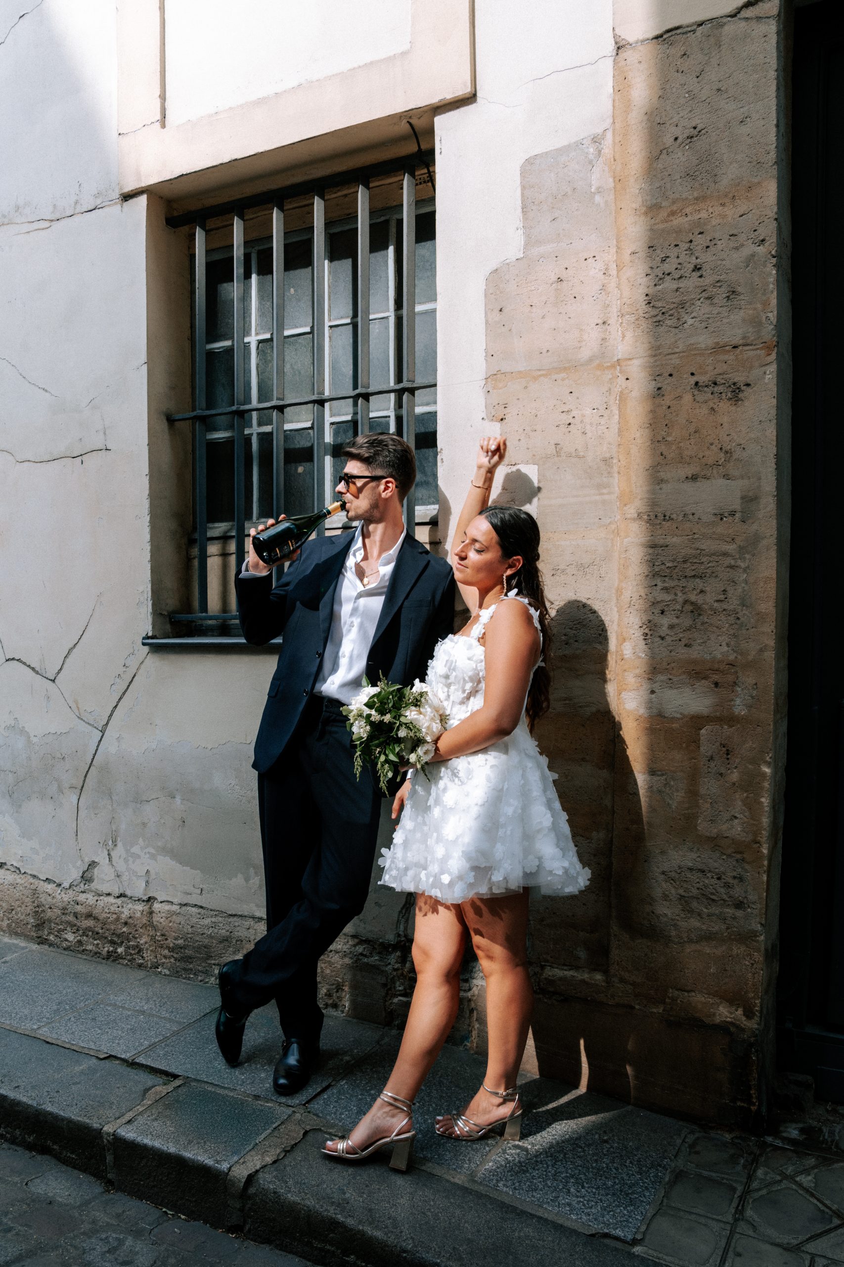 Un couple de jeunes mariés pose devant une façade ancienne aux murs patinés et à la fenêtre protégée par des barreaux. Le marié, vêtu d’un costume sombre, boit directement à une bouteille tandis que la mariée, en robe blanche fleurie, tient un bouquet et lève le bras dans une attitude festive.