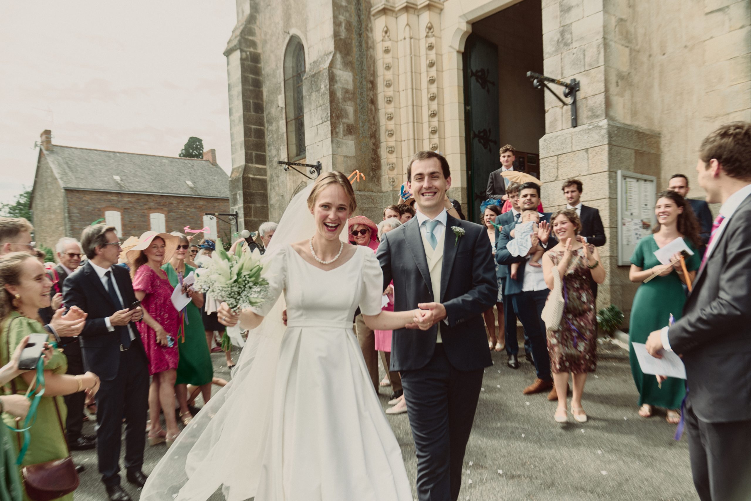 Une mariée en robe blanche et son époux en costume avancent joyeusement devant une église en pierre. Autour d’eux, leurs invités applaudissent, sourient et célèbrent leur sortie, certains tenant des fleurs, des rubans ou un bébé.
